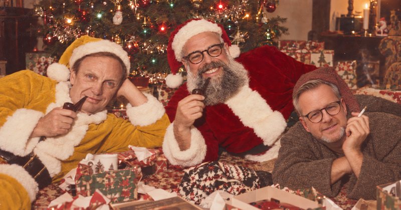 Tre tomtar i gul, röd och grå dräkt ligger framför en julgran: arbetsgivarjul, arbetarjul och gråsossejul.